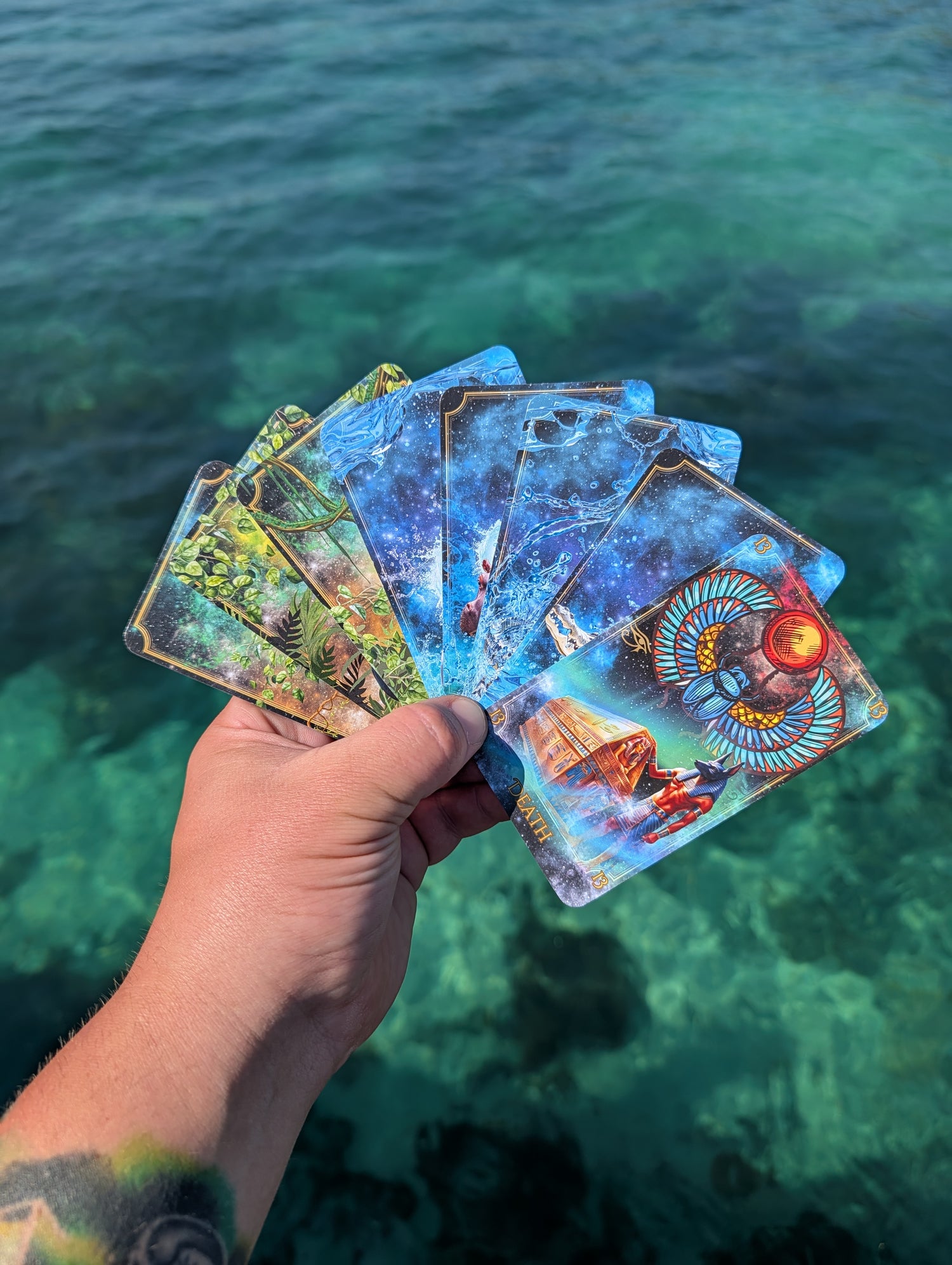 Written in the Stars Tarot cards by Emporos held above turquoise water — plastic-free tarot deck designed in Devon, printed in Cornwall, travels the world